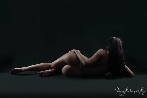 A naked brunette lies on a dark background with her back to the viewer