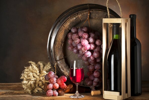 En la mesa, una botella en una Caja y una Copa de vino, y al lado en un barril, un racimo de uvas