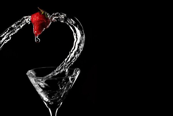 A glass on a black background into which a stream of water with strawberries pours