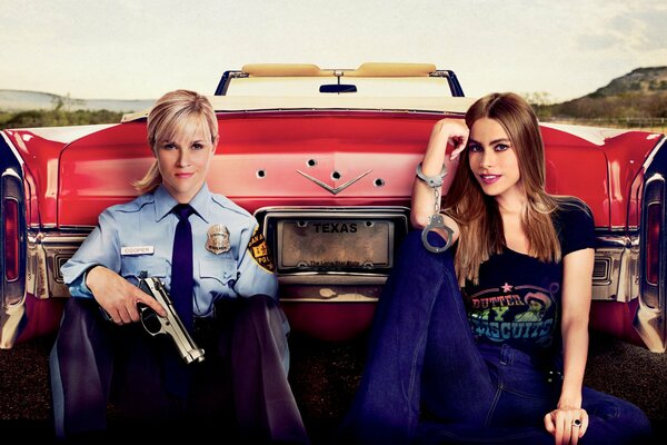 Against the background of a red car sits Reese Witherspoon in a police uniform with a gun and Sofia Vergara in handcuffs