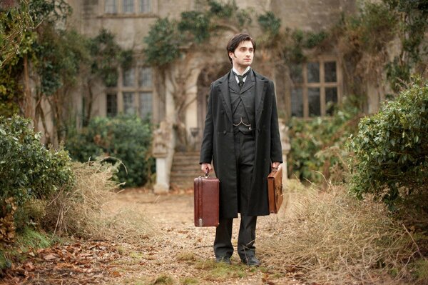 Daniel Radcliffe with suitcases in a black suit in front of an abandoned mansion