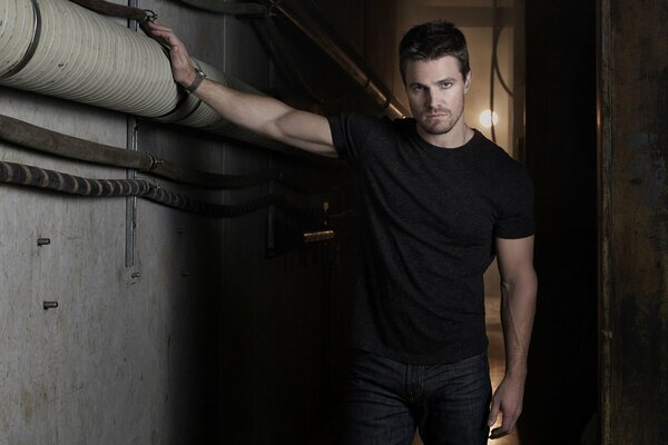 A man in a black T-shirt leans on a pipe from the TV series Arrow