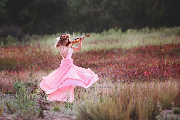 En el campo, una chica con un vestido rosa da vueltas y toca el violín