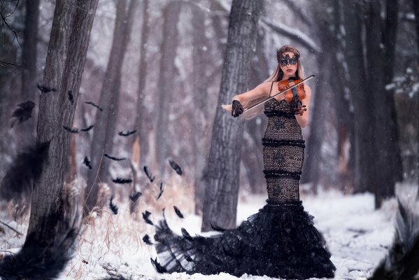 A girl in a mask and feathers plays the violin in a winter forest