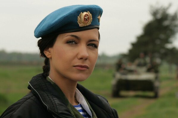 A beautiful woman in a military uniform looks at the viewer against the background of a tank leaving