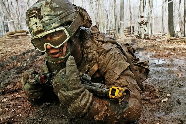 A soldier with a gun and in uniform is undergoing training, crawling on the muddy ground after the rain