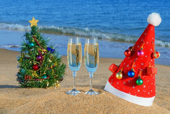 Рождественская ёлка, шапка Санты Клауса и два бокала шампанского на пляже океана