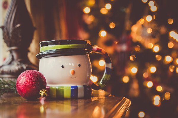 A snowman mug and a red Christmas ball on a camode against a background of lights