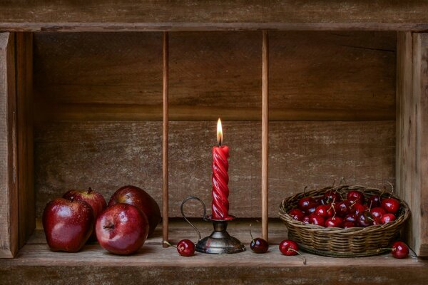 Eine rote verdrehte Kerze brennt zwischen Kirschen im Korb und Äpfeln auf einem Holzregal