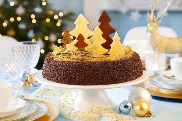 Рождественский самодельный торт со сладкими клочками на фоне сияющей новогодней красавицы ёлки оленя новогодних шаров и красивой белой посуды