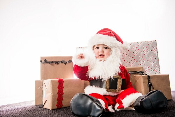 Ein kleines Kind im Weihnachtsmann-Kostüm sitzt mit Geschenken auf dem Hintergrund einer Krone und gratuliert allen zum neuen Jahr und zu Weihnachten