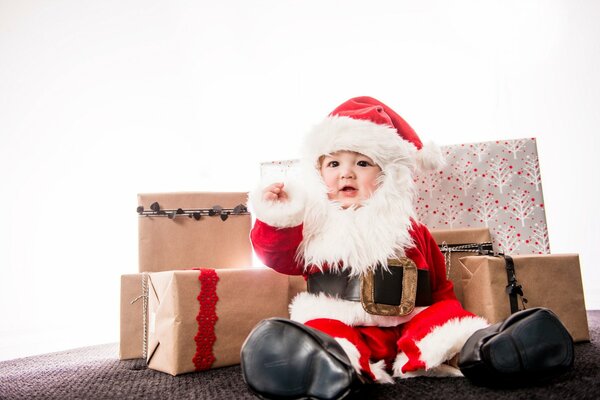 A small child in a Santa Claus costume is sitting on the background of a Coro ok with gifts and congratulates everyone on the New Year and Merry Christmas