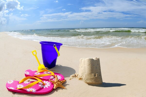 Pink slates with daisies, a blue bucket with a yellow scoop and a tower of sand against the background of the sea with a moving wave and a blue sky