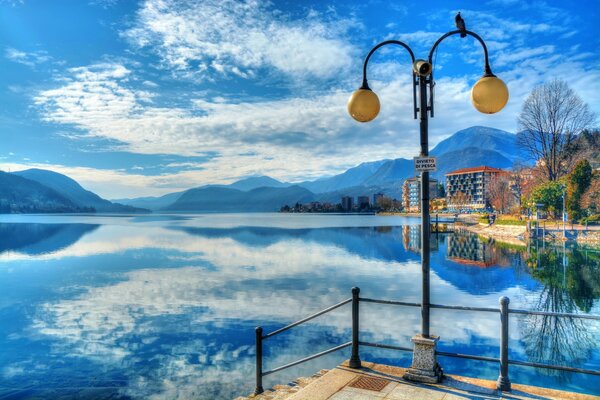 A lantern and houses on the shore of a blue lake in which clouds and mountains are reflected