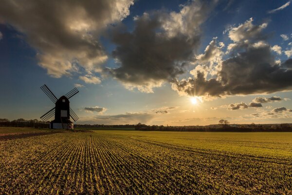 A mill in a vast field is illuminated by the sun breaking through thick clouds