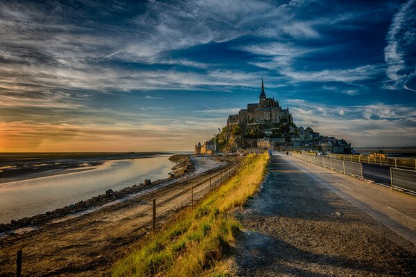 Zamek na wyspie Mont Saint Michel w promieniach wieczornego słońca pod błękitnym niebem z chmurami