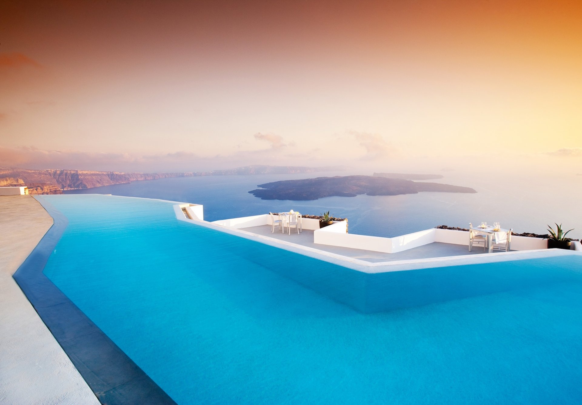 santorin grèce piscine hôtel tables fauteuils terrasse mer montagnes îles soirée coucher de soleil