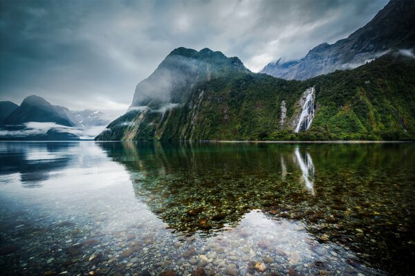 Neuseeland. Ein See, der von Bergen mit Wasserfällen umgeben ist, mit klarem Wasser, durch das der Boden sichtbar ist