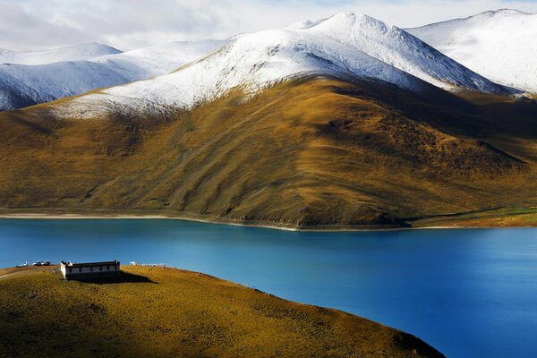 Maison solitaire au bord de la rivière sur fond de montagnes enneigées du Tibet