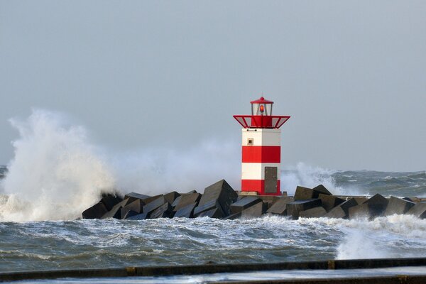 Die Meereswelle bricht an einem steinigen Untergrund mit einem rot-weißen Leuchtturm zusammen