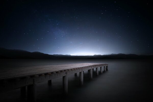 The starry sky above the pier near the lake. Mountains can be seen in the distance